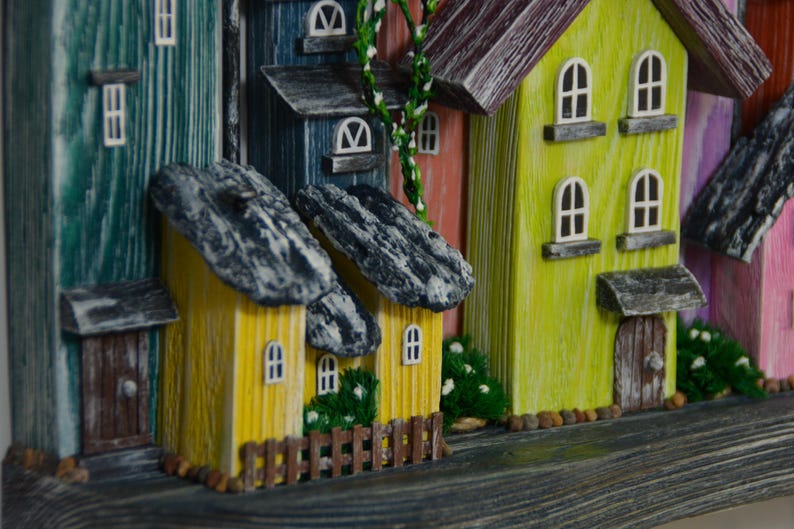 May include: A colorful, handcrafted miniature village scene. The houses are painted in various hues, including yellow, blue, and pink, with stone-like roofs. Small windows and doors add detail, and miniature greenery is placed around the houses.