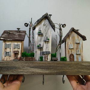 Può includere: Un portachiavi in legno con tre case in miniatura, ognuna con un design unico e dipinta in tonalità di marrone, bianco e blu. Le case sono fissate a una base in legno con ganci per appendere le chiavi.