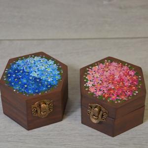 Może przedstawiać: Dwa sześciokątne drewniane pudełka z kwiatowymi wzorami. Jedno pudełko ma niebieski wzór kwiatowy, drugie różowy. Oba pudełka mają ciemnobrązowe wykończenie i mosiężne zapięcie.