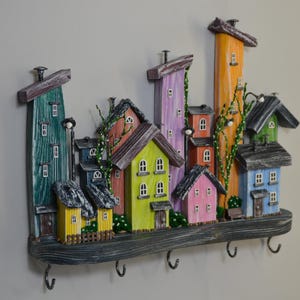 May include: A colorful wooden wall-mounted key holder featuring a whimsical village scene. The houses are painted in various colors, including green, yellow, pink, and blue, with dark gray roofs. The key holder has five metal hooks.