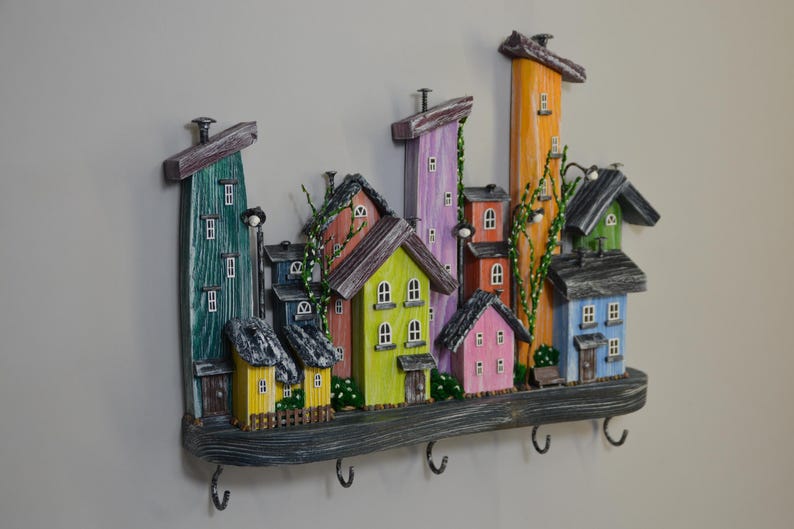 May include: A colorful wooden wall-mounted key holder featuring a whimsical village scene. The houses are painted in various colors, including green, yellow, pink, and blue, with dark gray roofs. The key holder has five metal hooks.