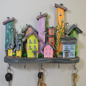 May include: A whimsical key holder featuring a miniature town scene with colorful houses and buildings. The houses are painted in various colors, including green, yellow, pink, and blue. The key holder has six hooks and is mounted on a wooden base.