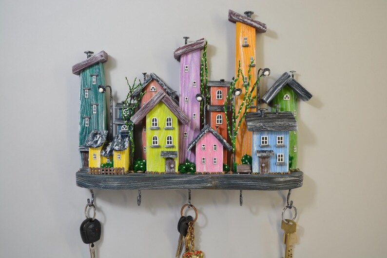 May include: A whimsical key holder featuring a miniature town scene with colorful houses and buildings. The houses are painted in various colors, including green, yellow, pink, and blue. The key holder has six hooks and is mounted on a wooden base.