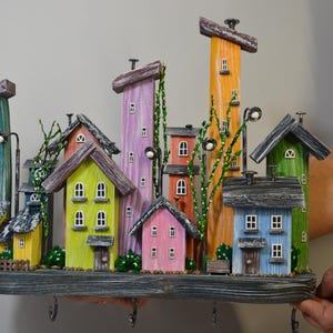 May include: A colorful, whimsical key holder featuring a miniature village scene. The houses are painted in various bright colors, including green, yellow, pink, and blue, with dark gray roofs. The key holder has several hooks and decorative street lamps.