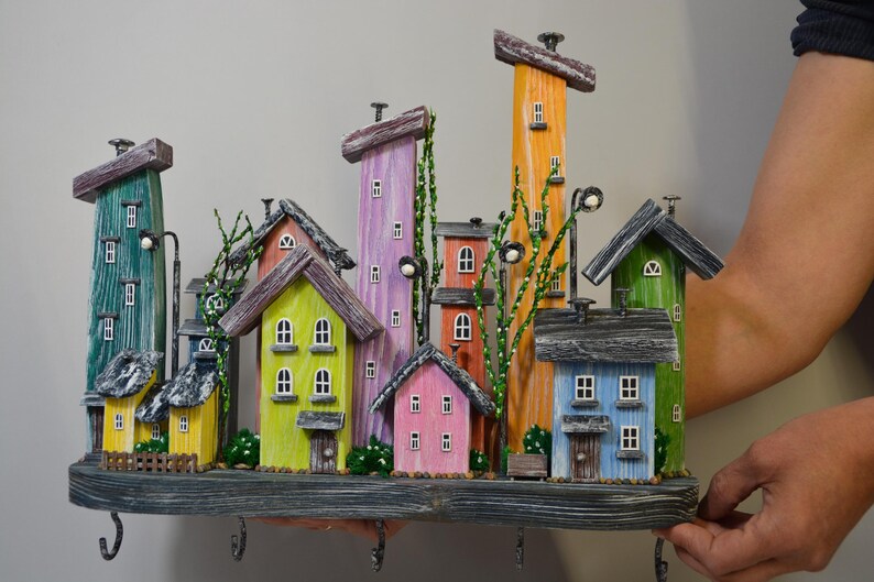 May include: A colorful, whimsical key holder featuring a miniature village scene. The houses are painted in various bright colors, including green, yellow, pink, and blue, with dark gray roofs. The key holder has several hooks and decorative street lamps.