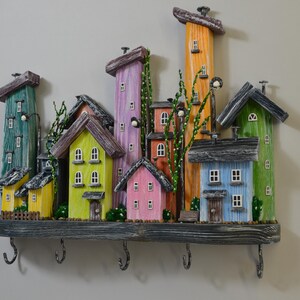 May include: A colorful wooden key holder shaped like a whimsical village. The houses are painted in various colors, including green, yellow, pink, orange, and blue. The key holder has several hooks and is mounted on a gray wooden base.