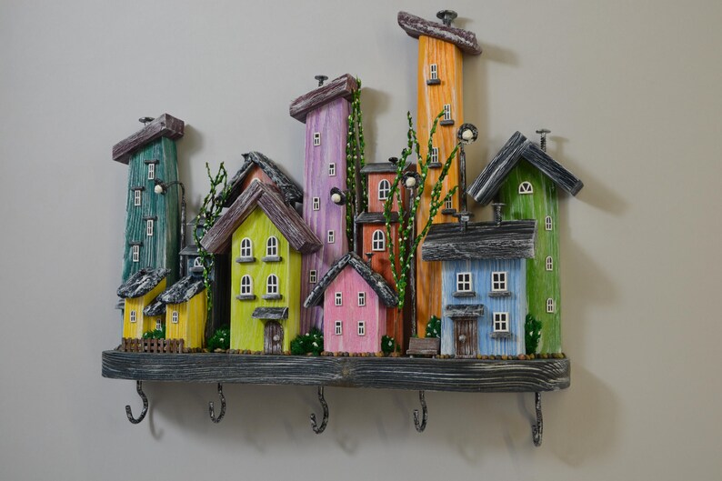 May include: A colorful wooden key holder shaped like a whimsical village. The houses are painted in various colors, including green, yellow, pink, orange, and blue. The key holder has several hooks and is mounted on a gray wooden base.