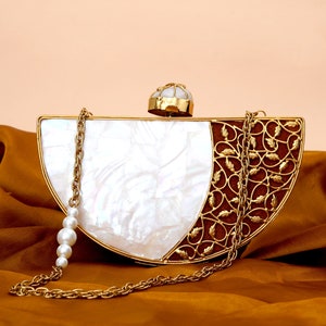 Puede incluir: Un clutch de media luna blanco y dorado con una correa de cadena de perlas. El clutch tiene un diseño metálico decorativo en un lado y un acabado de nácar blanco en el otro.