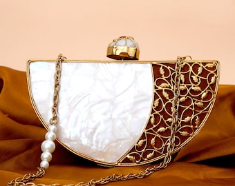 May include: A white and gold half-moon shaped clutch with a pearl chain strap. The clutch has a decorative metal design on one side and a white mother-of-pearl finish on the other side.