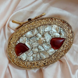 Puede incluir: Un embrague ovalado dorado con un borde decorativo. El embrague está adornado con un mosaico de piezas de concha blanca e iridiscente, acentuado con gemas rojas y pequeñas perlas blancas.