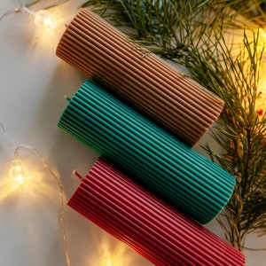 Op de afbeelding: Drie geribbelde pilaar kaarsen in bruin, teal en rood, diagonaal gerangschikt. De kaarsen zijn cilindrisch met een textuur. Groene dennentakken en een lichtsnoer met warmwitte lampen op de achtergrond.