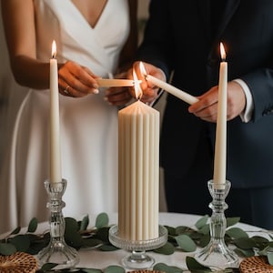 May include: A central ribbed pillar candle on a glass pedestal, flanked by two tall taper candles in clear glass holders. The candles are lit, with flames visible. Green eucalyptus leaves and round woven coasters are arranged on the table.