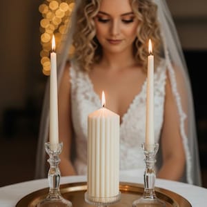 Puede incluir: Una escena de boda con tres velas encendidas sobre una bandeja dorada. Dos velas altas y delgadas en portavelas de vidrio transparente flanquean una vela de pilar más grande y acanalada. Las velas se colocan sobre un fondo borroso.