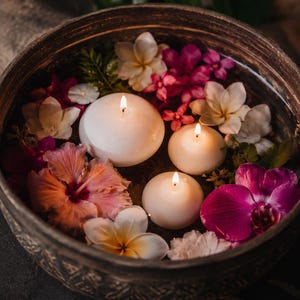 Peut inclure: Un bol circulaire sombre et texturé rempli d'eau, contenant trois bougies flottantes blanches rondes aux mèches allumées, ainsi qu'un assortiment de fleurs colorées, dont des hibiscus roses, des orchidées violettes et des fleurs de frangipanier.