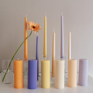 Puede incluir: Una colección de velas coloridas, incluyendo velas de pilar y cónicas. Las velas de pilar tienen ranuras verticales y vienen en tonos naranja, morado y amarillo. También hay velas cónicas en tonos similares, con una flor de gerbera.