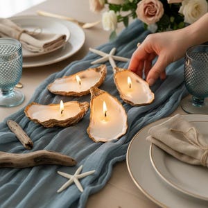 Könnte beinhalten: Vier brennende Austernschalen-Kerzen mit goldenem Rand liegen auf einem blauen Tischläufer. Der Tisch ist mit Tellern, Gläsern und Blumen gedeckt. Treibholz und Seesterne ergänzen das Küsten-Thema.