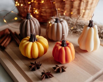May include: Five decorative pumpkin-shaped candles on a wooden surface. The candles are in shades of brown, beige, yellow, and orange. There are also cinnamon sticks and star anise on the surface.