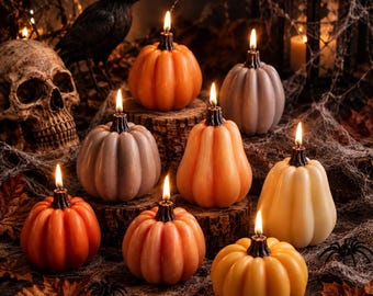 May include: A collection of eight pumpkin-shaped candles in shades of orange, cream, and grey, each featuring a lit wick with a dark base. The candles are arranged among dried autumn leaves, a human skull, a black raven, and spider webs.