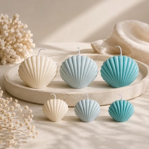Peut inclure: Un ensemble de six bougies en forme de coquillage disposées sur deux rangées sur un plateau circulaire. Les bougies sont de trois couleurs : crème, bleu clair et turquoise. Chaque bougie présente une texture côtelée et une mèche blanche.