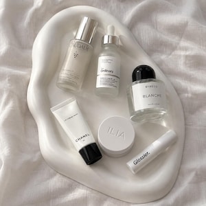 Può includere: Una collezione di prodotti per la cura della pelle e profumi che include un siero Caudalie, un flacone contagocce The Ordinary, profumo Byredo Blanche, crema mani Chanel La Crème Main, un vasetto Ilia e un tubetto Glossier su un vassoio bianco.