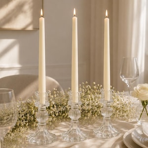 Peut inclure: Trois bougeoirs en verre transparent avec des tiges nervurées et des bases larges, chacun portant une bougie effilée longue et fine de couleur blanc cassé avec une flamme allumée. Ils sont disposés en rangée avec des petites fleurs blanches.