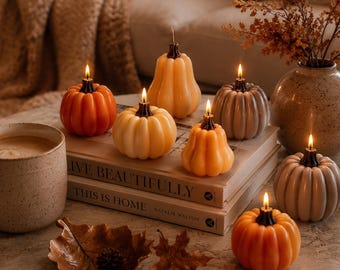 May include: A collection of six pumpkin-shaped candles in shades of orange, cream, and grey, each with a lit wick. The candles are arranged on two stacked books titled LIVE BEAUTIFULLY and THIS IS HOME, alongside a ceramic mug and a leaf-shaped dish.
