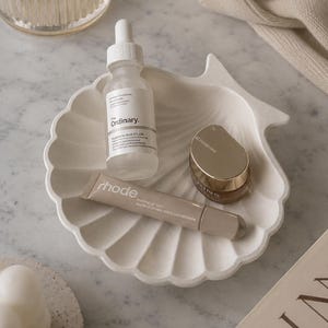 May include: A white, shell-shaped ceramic tray holds three skincare products: a clear glass dropper bottle labeled The Ordinary Hyaluronic Acid 2% + B5, a beige tube labeled rhode Peptide Lip Tint, and a small brown jar with a gold lid labeled Clarins.