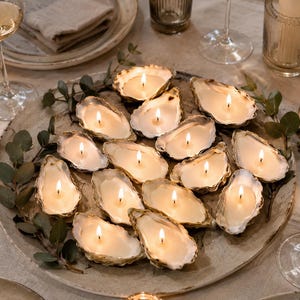 Peut inclure: Une collection de quatorze bougies en coquille d'huître avec des mèches allumées, disposées sur un grand plateau en céramique texturée. Les bougies présentent de la cire blanc cassé coulée dans des coquilles d'huîtres naturelles et irrégulières.