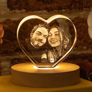 May include: A heart-shaped crystal photo, lit from within, displaying a couple's portrait. It rests on a wooden base, placed on a yellow box. The background features a brick wall and a gramophone.