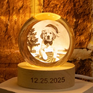 May include: A lit glass sphere with a detailed image of a dog in a Santa hat and bow tie, with a Christmas tree. The sphere sits on a wooden base engraved with "12.25.2025". The base rests on a white, hexagonal platform. The light has a warm glow.
