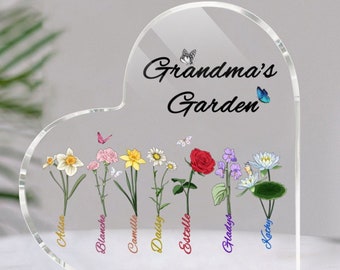 May include: A heart-shaped clear acrylic plaque with the text "Grandma's Garden" and a butterfly design. Seven different flowers are displayed with names below each flower: Alissa, Blanche, Camille, Daisy, Estelle, Gladys, and Kathy.