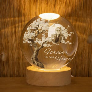 Può includere: Una sfera di vetro trasparente con una foto e un disegno di albero, illuminata da una luce calda. La sfera poggia su una base di legno, con la scritta "Forever in our Heart" incisa sul davanti.