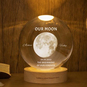 Puede incluir: Una esfera de cristal transparente con una imagen de la luna y las palabras "OUR MOON". Los nombres "Aimee" y "Vicky" están a ambos lados de la imagen de la luna. La fecha "04.05.2023" y la frase "THE BEGINNING OF OUR FOREVER" están debajo. La esfera se apoya en una base de madera.