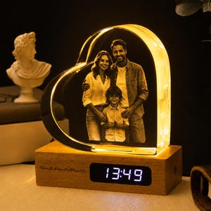 Könnte beinhalten: Eine herzförmige, klare Acrylplatte mit einem schwarz-weißen Foto einer dreiköpfigen Familie, montiert auf einem rechteckigen Holzsockel mit einer digitalen Uhranzeige, die 13:49 Uhr und den Text Home Is Where We Are anzeigt.