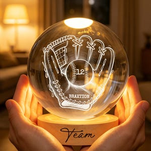 Könnte beinhalten: Eine klare, kugelförmige Glaslampe mit einer Gravur eines Baseballhandschuhs und Balls, der Nummer 12 und dem Namen BRAXTION. Die Kugel ruht auf einem runden Holzsockel, in den das Wort Team eingraviert ist und der ein warmes Licht ausstrahlt.