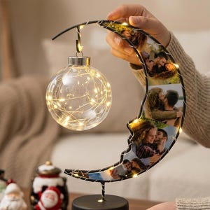 May include: A crescent moon-shaped lamp featuring multiple printed photographs along its frame. A clear glass sphere filled with warm LED fairy lights hangs from the top of the moon. The entire structure is supported by a solid black circular base.