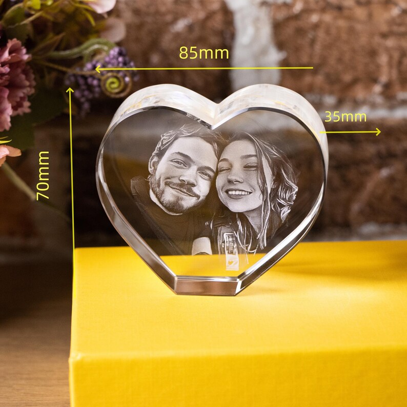 May include: A clear, heart-shaped crystal photo block, measuring 7cm high, 8.5cm wide, and 3.5cm deep, displaying a black and white image of a smiling couple. The crystal is set on a yellow surface, with flowers in the background.