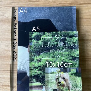 May include: Two acrylic photo frames with a black and white photo of a person in a black shirt and a photo of a child in a green field. The smaller frame is 10x10cm and has the text "I LOVE RUNNING with Daddy!" in white. The larger frame is A4 size and has the text "Happy Father's Day 2023" in white.
