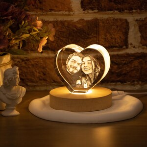 May include: A heart-shaped crystal photo featuring a couple, lit by a warm glow. The crystal sits on a wooden base, placed on a white, irregular-shaped surface. The background includes a brick wall and decorative items.