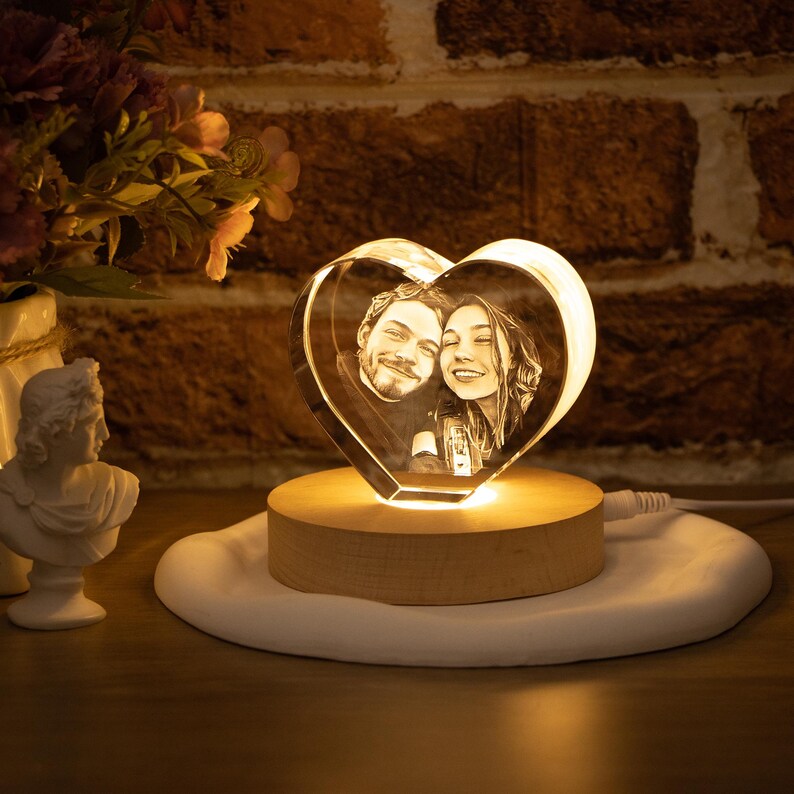 May include: A heart-shaped crystal photo featuring a couple, lit by a warm glow. The crystal sits on a wooden base, placed on a white, irregular-shaped surface. The background includes a brick wall and decorative items.
