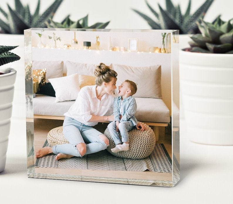 May include: A mother and child are sitting on a floor with a light gray rug. The mother is wearing a white shirt and blue jeans. The child is wearing a blue shirt and blue jeans. They are both smiling and looking at each other.