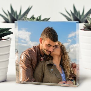May include: A couple in brown leather jackets, the woman is kissing the man on the cheek, against a blue sky with white clouds.