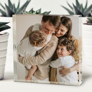 May include: A family of four, a man, a woman, and two children, are hugging each other. The man is wearing a beige shirt, the woman is wearing a yellow floral dress, the girl is wearing a white dress, and the boy is wearing a white shirt. The family is smiling and looking at each other.
