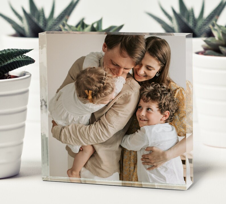 May include: A family of four, a man, a woman, and two children, are hugging each other. The man is wearing a beige shirt, the woman is wearing a yellow floral dress, the girl is wearing a white dress, and the boy is wearing a white shirt. The family is smiling and looking at each other.