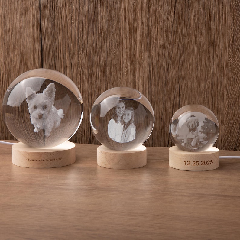May include: Three illuminated glass spheres with engraved images of a dog, a mother and daughter, and a dog with a person. Each sphere sits on a wooden base. The largest sphere has the text "Love is a her biggest word".
