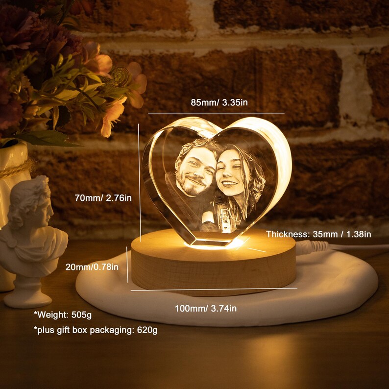 May include: A heart-shaped crystal photo featuring a couple, lit by a warm glow. The crystal is 8.5cm wide, 7cm high, and 3.5cm thick, set on a wooden base. The base sits on a white circular platform. The weight is 505g.