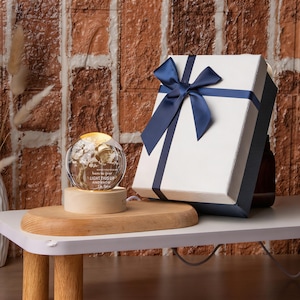 May include: A lighted glass sphere with a floral design and the text "LIGHT THIS UP AND KNOW". It sits on a wooden base. A white gift box with a navy blue ribbon is next to it. The background is a brick wall.