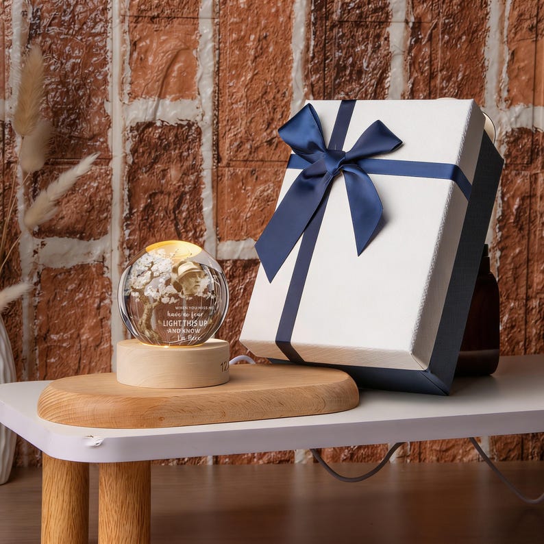 May include: A lighted glass sphere with a floral design and the text "LIGHT THIS UP AND KNOW". It sits on a wooden base. A white gift box with a navy blue ribbon is next to it. The background is a brick wall.