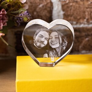 May include: A clear, heart-shaped crystal photo frame displaying a black and white image of a smiling couple. The frame sits on a yellow surface, with flowers and a brick wall in the background.