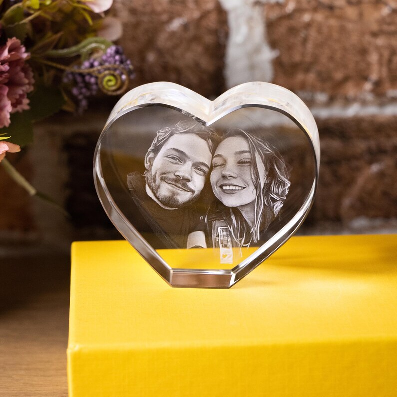 May include: A clear, heart-shaped crystal photo frame displaying a black and white image of a smiling couple. The frame sits on a yellow surface, with flowers and a brick wall in the background.
